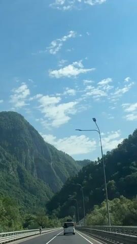 A vertical, first-person perspective video shows a highway carved into a lush, green mountain valley under a bright blue sky with scattered white clouds. A white van is traveling on the highway in the same direction as the camera. A street lamp with a single light fixture is visible on the right side of the road, and another further down the road. The mountains are covered in dense green foliage.