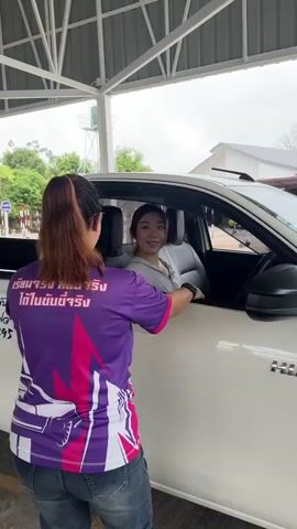 The video shows a woman getting out of a white pickup truck under a metal roof structure, likely at a driver training facility. A woman in a purple shirt with text on the back helps her exit. The camera pans to show a man wearing a green safety vest with the number "16" on it, standing near the truck. Other people, also wearing numbered vests, are walking or riding motorcycles in the background. The environment appears to be an open-air covered area with driving course markers visible. The scene depicts a practical driving test or training session.