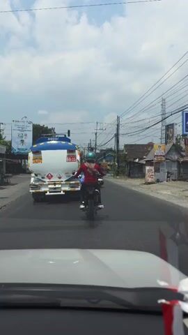 A rear view of a motorcycle rider on a road. The rider is wearing a red shirt, blue pants, and a teal helmet. The motorcycle is traveling away from the camera. Behind the motorcycle is a white and blue tanker truck with the number "60" visible on its side. The road is paved and appears to be in a rural or suburban area. There are power lines and some buildings visible along the side of the road. The sky is cloudy. The camera appears to be positioned inside a vehicle, looking forward.