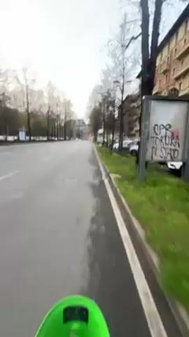 The video starts with a view of a light-colored building and trees against an overcast sky. A young man wearing a hooded jacket is visible briefly before the camera shifts to show a green electric scooter moving down a city street. The street is paved, with a sidewalk and trees lining the side. Cars are parked along the sidewalk. The video continues to show the view from the scooter as it travels down the street, with buildings and more trees visible in the background. The sky remains overcast.
