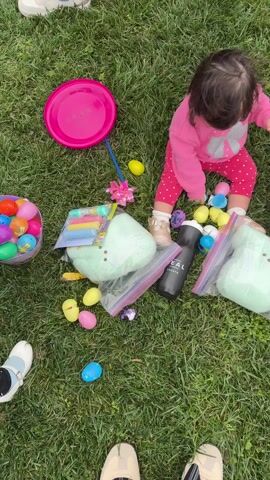 The video shows a toddler seated on a grassy lawn surrounded by Easter-themed items. She's wearing a pink outfit and is reaching for colorful plastic Easter eggs scattered around her. There are two purple Easter baskets filled with eggs, two clear plastic bags containing light green plush bunny-shaped items, a pink frisbee, and a black water bottle labeled "REAL CHURCH". Adults and children are scattered in the background across the lawn, participating in an Easter egg hunt. The event appears to be taking place outdoors in a park-like setting with bare trees visible in the distance.