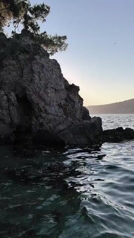 The video shows a body of water with waves under a clear sky. The camera pans from a rocky shoreline with a few trees to a distant landmass with hills and buildings. The water appears dark and reflects the sky and surrounding landscape. The overall scene suggests a natural coastal environment.