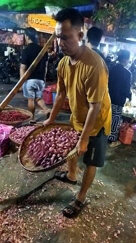 A person wearing a yellow t-shirt and black shorts is standing in an outdoor market at night. They are holding a woven basket and shaking it, causing its contents to spill. The basket appears to be filled with small, pinkish-purple items, possibly dried flowers or spices. In front of the person, there are other baskets also containing similar items. The ground is covered with scattered pinkish-purple debris. In the background, there are stalls with merchandise, including a sign that says "INDOMIE." Other people are visible in the background, suggesting a busy marketplace. The lighting is dim, typical of an evening market.