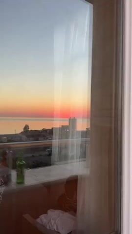 The video opens with a close-up reflection of a sunset over the ocean on a glass pane. The reflection shows a gradient of orange, pink, and yellow hues in the sky above the water. In the foreground of the reflection, a blurry cityscape with some tall buildings is visible. The camera then pans to the right, revealing the interior of a room with unfinished walls. The walls are partially covered with grey plaster and some orange-colored planks. Next, the camera moves to an exterior view from a balcony, showcasing a panoramic landscape. A metal railing runs across the bottom of the frame. Beyond the railing, a road with several trucks is visible. To the left of the road, there are buildings and a green, hilly landscape. To the right of the road, the coastline stretches out, with the ocean meeting the horizon. The sunset is still visible, casting a warm glow over the scene. The sky is clear and transitions from a light blue at the top to shades of orange and pink near the horizon. Several industrial structures, including silos and what appears to be a construction site, are present near the coast. The video ends by focusing on the sunset over the ocean, with more trucks visible on the road in the foreground.