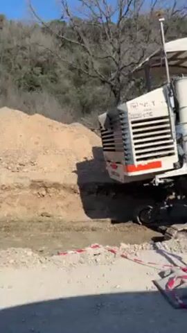 The video shows a large, tracked machine with a white and orange color scheme, identified as a Wirtgen, working on a construction site. The machine is positioned on a dirt road, cutting into a dirt bank. An operator wearing a safety vest is visible on the machine. A red and white striped traffic barrier and a yellow warning sign are positioned along the side of the road. The surrounding environment includes trees and a clear blue sky.