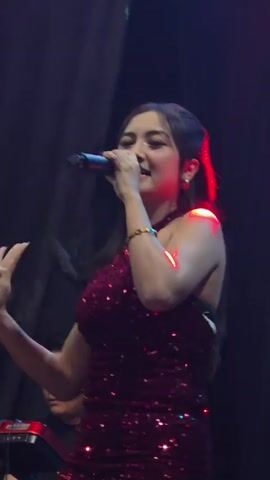 A woman in a sparkling red dress sings into a microphone. She has long, dark hair and is wearing gold earrings and a bracelet. Behind her, a man plays a keyboard. The stage lighting is a mix of red and purple hues.