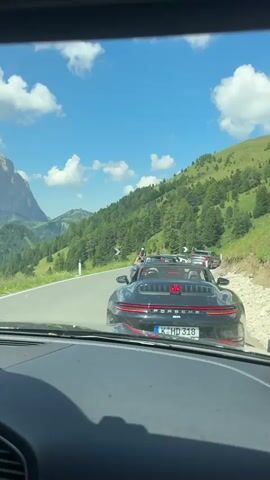 The video shows a view from inside a car driving on a mountain road. In the foreground, the dashboard of the car is visible, including a screen displaying a rear-view camera feed. Outside the car, a black Porsche convertible with red taillights is driving ahead. The road winds through a mountainous landscape with lush green hillsides dotted with pine trees. In the background, imposing rock formations rise dramatically against a bright blue sky with scattered white clouds. The scene suggests a scenic drive through the Alps.