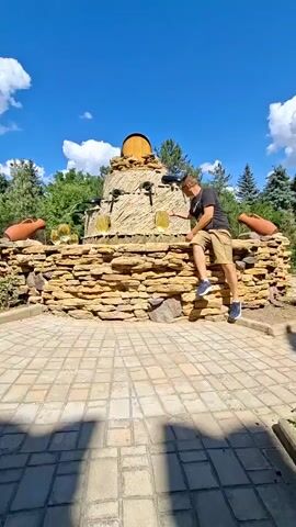 The video shows a man in a black t-shirt and beige shorts standing on a stone fountain. He is reaching out to touch a part of the fountain. The fountain is made of stacked stones with decorative amphorae on either side. In the background, there are trees and a blue sky with clouds. The ground in front of the fountain is paved with interlocking bricks. The man appears to be in good health. The scene suggests an outdoor recreational area.