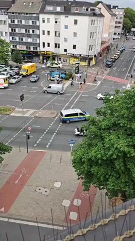 The video shows a street intersection from an elevated perspective. Several vehicles are visible, including a yellow delivery van, a white passenger car, a white minivan, a black sedan, and a police van. Pedestrians are also present, crossing the street and walking on the sidewalks. The intersection appears to be busy with traffic. Buildings line the street, some of which have storefronts. Trees are present on the sidewalks and near the intersection. The sky is overcast.