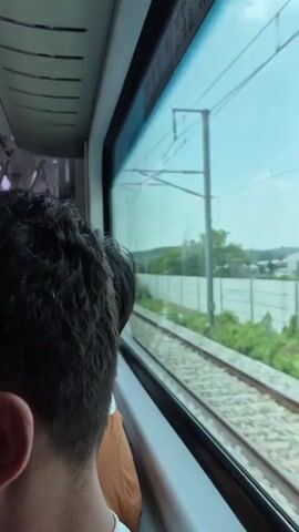 The video shows the interior of a train with passengers. In the foreground, a person's head and shoulder are visible from behind, looking out a window. Outside the window, train tracks and greenery are visible. The camera then pans to show other passengers inside the train. One man in a white shirt is facing away from the camera, and another man in a grey shirt is standing by the window. The train appears to be in motion.