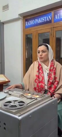 A woman wearing a white headscarf and a light brown shawl with red floral designs is sitting in front of a vintage reel-to-reel audio recorder in what appears to be a Radio Pakistan studio. The recorder is gray with multiple knobs and buttons, and a reel of tape is visible. Behind her is a glass-fronted cabinet with the words "RADIO PAKISTAN AUDIO" displayed above it in blue lettering. The room seems to be well-lit, and there is a table with papers to the left of the woman. She is looking to the right and speaking.