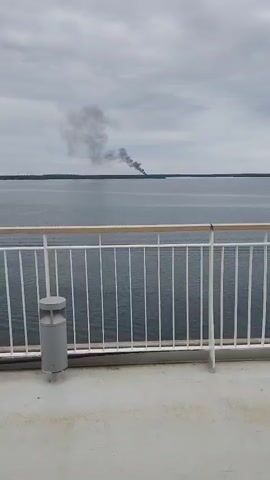 The video shows a body of water under a cloudy sky. In the distance, a plume of black smoke rises from a forested shoreline. The foreground of the video features a white railing and a grey trash receptacle. A white speaker is attached to the railing. The viewpoint is from a deck, likely on a vessel.