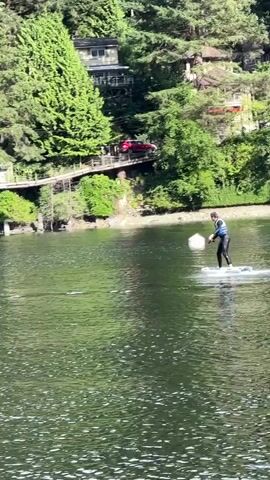 A person wearing a black wetsuit and a blue life vest is riding a white electric hydrofoil board on calm, dark water. The person is standing on the board, facing the right side of the frame, and appears to be in motion, leaving a small wake. The background shows a densely forested shoreline with several houses built on stilts or a hillside. A red car is visible parked on a driveway leading up to one of the houses. In the water, to the right of the person, there is a white buoy. The weather appears to be sunny, casting reflections on the water.