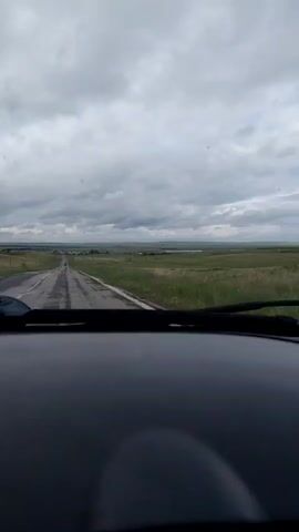 A view from inside a vehicle, looking out through the windshield at a straight, paved road that recedes into the distance. The road is flanked by green fields on either side. The sky is overcast with heavy, gray clouds. The dashboard of the vehicle is visible in the foreground, along with the windshield wipers. The overall impression is a journey through a somewhat somber, open landscape.