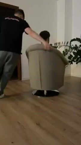 Two men are in a room with wood flooring. One man sits in a gray swivel chair while the other pushes the chair. The first man falls over backwards in the chair. There is a large green plant visible in the background. There is also a door on the left of the screen.