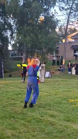 A group of people are performing on a grassy lawn in front of a building. A woman in a blue jumpsuit and red accents is dancing in the foreground. Other performers in various costumes are scattered around the lawn. Trees and a building are visible in the background.
