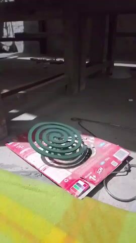 The video shows a mosquito coil burning. The coil is green and spiral-shaped, and it is placed on top of a red and white cardboard box. The box appears to be a product package for the mosquito coil. Smoke is rising from the burning coil, indicating that it is actively producing insecticide. The background is dimly lit and appears to be an indoor or sheltered outdoor space, possibly a shed or a covered patio, with wooden structures visible above and a concrete floor below. The overall scene suggests a practical application of the mosquito coil for pest control.