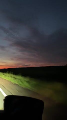 A car is driving down a highway at dusk. The sky is a deep blue with streaks of orange and pink from the setting sun. The headlights of the car illuminate the road and the grassy shoulder. A side mirror of the car is visible in the foreground. The landscape is flat and dark, with a line of trees in the distance. The video is taken from the passenger seat of the car.