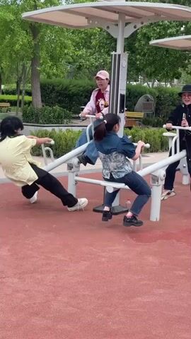 Here is a description of the video:

The video shows four people on a playground apparatus that looks like a series of seesaws connected to a central tower with a roof. The apparatus is grey and white and set on a red playground surface. The people are all sitting on the seats and working the seesaw motion. The person on the left wears a yellow shirt and black pants. Next to them, a person with a ponytail wears a blue top and blue jeans. On the third seesaw, a person with a pink hat and pink jacket also wears blue jeans. The person on the right wears a black jacket and black pants. Behind the apparatus, there are green trees and benches in the park.