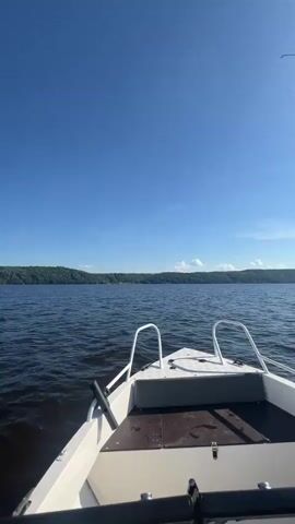 The video shows a first-person view from a boat moving on a body of water. The boat is white with chrome railings. The water is dark and choppy, creating a wake behind the boat. The sky is a vibrant blue with scattered white clouds, indicating a clear and sunny day.

In the distance, a long, tree-covered shoreline is visible. The trees are dense and green, suggesting a natural, possibly remote, landscape. The sun is high in the sky, casting a bright light and a slight glare on the water.

The overall mood of the video is peaceful and adventurous, conveying the experience of boating on a pleasant day. The absence of people suggests a solitary or private outing. The visual elements combine to create a serene and natural scene, characteristic of a summer day spent on the water. The location is identified as Siukterka, Russia.