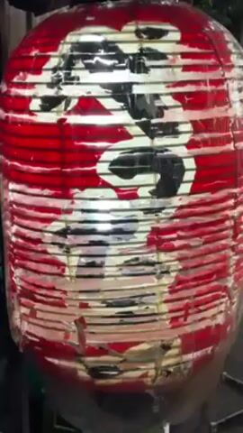A close-up shot of a traditional Japanese paper lantern, predominantly red and white with black Kanji characters, is presented in the video. The lantern's surface is worn and partially peeling, revealing the light source within. The lantern is cylindrical in shape, with horizontal red stripes alternating with white sections where the characters are painted. The bottom of the lantern appears damp or slightly damaged, suggesting exposure to the elements. The background is dark and indistinct, implying an outdoor setting at night or in low light. The camera remains relatively static, keeping the lantern in focus throughout the short video.