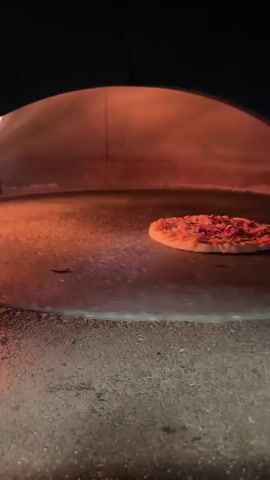 A single pizza sits on the floor of a brick oven. The oven is glowing with heat, and flames can be seen in the background. The camera is positioned low, looking up into the oven at the pizza. The video is shot in a way that makes the pizza appear to be the sole focus, with the environment surrounding it rendered in a warm, inviting glow. The pizza appears to be cooking, with some browning on the crust and melted cheese visible. There are no people or other objects in the frame.
