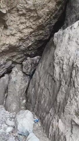 The video shows a dark, narrow cave entrance with rocky walls. The ground is covered in loose stones and dirt. The camera slowly pans across the opening, revealing the darkness within.