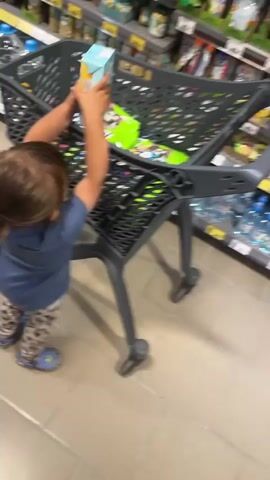 A young child with dark hair pulled into a ponytail, wearing a blue t-shirt and leopard-print leggings, stands at the side of a dark gray shopping cart in a supermarket aisle. The child reaches into the cart, where several items are visible, including a large green bag with a cartoon dog on it and a light blue box. The child's left hand is raised slightly above the cart. The shopping cart has a handle that a person's hand is gripping from the first-person perspective. In the background, shelves stocked with various products line the aisle, illuminated by overhead lighting. The floor is tiled. The video appears to be taken from a slightly elevated angle, looking down at the child and the cart.