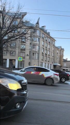 The video shows a dark-colored SUV driving down a city street with light-colored buildings on either side. The SUV is the primary focus and occupies a significant portion of the frame. The buildings vary in architectural style, with some being taller and more modern, and others appearing older with distinct features. Power lines are visible in the sky. Other vehicles are present on the road, including a white SUV in the distance. The sky is blue and mostly clear, suggesting a sunny day.  The video seems to be taken from a moving vehicle.