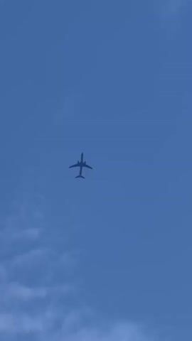 A drone perspective video shows an airplane flying in a clear blue sky. The airplane is silhouetted against the sky, and its details are not clearly visible. The airplane appears to be a commercial jet with two engines mounted under its wings. The airplane is flying at a high altitude, and it is moving from left to right in the frame. The video is shot from below the airplane, looking up at it. The airplane is the only object in the sky, and there are no other visible objects in the frame. The video is a continuous shot, and there is no editing or special effects. The video is 3 seconds long.