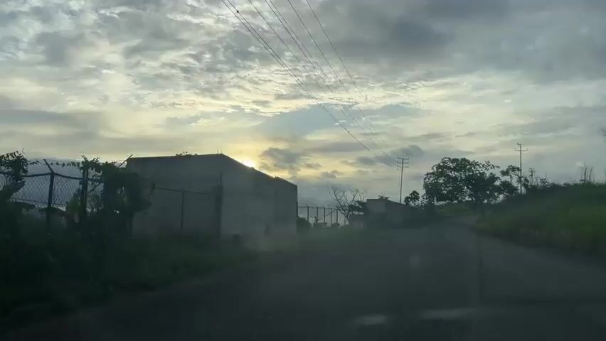 The video shows a view from a vehicle driving on a paved road. The road curves to the right. On the left side of the road is a tall concrete wall, and beyond that, a chain-link fence with barbed wire. Some trees are visible behind the fence. On the right side of the road, there is grass and a few trees, including a large tree with dense foliage. In the distance, there are utility poles with power lines strung between them. The sky is cloudy with some patches of sunlight breaking through, suggesting it is either sunrise or sunset. The overall environment appears to be semi-rural or industrial.