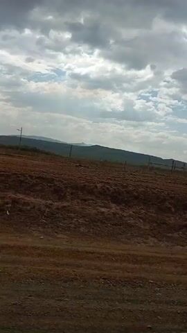 The video shows a landscape with a brown, mostly barren field in the foreground. There are a few scattered trees visible in the middle ground. In the background, there are low, rolling hills or mountains. The sky is overcast with patches of blue sky visible between the clouds. The camera appears to be moving, possibly from a vehicle. There is music playing in the background.