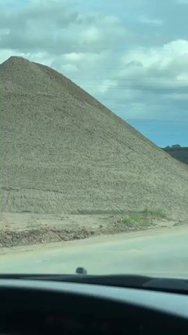 The video, shot from inside a vehicle, shows a large pile of light brown aggregate (likely sand or dirt) dominating the left side of the frame. The sky is cloudy with patches of blue, and power lines are visible across the upper middle of the frame. A paved road is in the foreground.

Around the 0:01 mark, a white and grey dump truck, with the brand "SHACMAN" visible on its side, begins to emerge from behind the large pile on the right. As the truck moves further into view, a rearview mirror becomes visible in the top left corner of the frame. This mirror reflects a yellow piece of heavy machinery, possibly an excavator, with what appears to be "CAT" (Caterpillar) text visible on it. The truck continues to slowly move or is parked behind the pile for the remainder of the clip. The scene suggests a construction or industrial site.