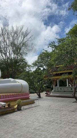 A panning video shows a large, white Buddha statue in a seated position. The statue is adorned with gold accents and sits on a multi-colored base. In front of the statue, a small offering table holds various items. To the left of the Buddha, a smaller golden statue is visible in front of a traditional temple structure with ornate golden roofs. The background consists of lush green trees and a partly cloudy blue sky. A few small potted plants are placed around the base of the large Buddha.