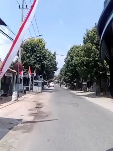 THE VIDEO SHOWS A VIEW FROM A MOTORCYCLE OR SIMILAR VEHICLE MOVING DOWN A TWO-LANE ROAD. THE ROAD IS PAVED AND APPEARS TO BE IN A SEMI-URBAN OR RURAL AREA, WITH BUILDINGS LINING BOTH SIDES. TREES ARE VISIBLE ON EITHER SIDE OF THE ROAD. SEVERAL RED AND WHITE FLAGS, CONSISTENT WITH THE INDONESIAN FLAG, ARE HUNG FROM POLES AND BUILDINGS. THERE ARE SEVERAL VEHICLES ON THE ROAD AHEAD, INCLUDING CARS AND WHAT APPEARS TO BE A TRUCK ON THE LEFT SIDE. THERE ARE ALSO PEOPLE VISIBLE, BUT THEIR FEATURES ARE NOT CLEAR DUE TO THE DISTANCE AND MOVEMENT. THE SKY IS CLEAR AND BLUE, INDICATING A SUNNY DAY. THE CAMERA IS FOCUSED STRAIGHT AHEAD, GIVING A FIRST-PERSON PERSPECTIVE OF THE JOURNEY.
