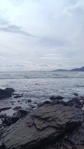 The video shows an ocean scene. A rocky shoreline, with a large, dark gray rock in the foreground, leads to the ocean. The water is choppy with waves breaking. In the distance, mountains are visible on the horizon under a cloudy, overcast sky. Sounds of people talking are heard in the background.