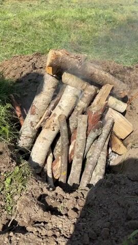 The video shows a pile of logs burning in a pit dug into the ground. The logs are of various sizes and types, with some appearing to be thicker branches and others smaller twigs. Smoke is rising from the pile, indicating that it is actively burning. The pit is surrounded by loose soil, and the ground around the pit is covered in grass. The lighting suggests it is daytime, possibly outdoors in a rural or natural setting. There are no people visible in the video. The focus is on the burning wood.