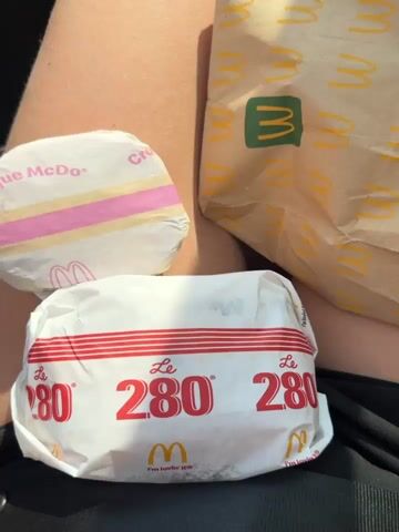 A person is sitting in a car. On their lap are two McDonald's hamburgers wrapped in branded paper, and a McDonald's paper bag. The hamburger in the foreground has "Le 280" printed on its wrapper. The hamburger behind it has a pink and white striped wrapper that says "Croque McDo." The McDonald's bag is a light brown color with yellow arches. The camera then pans to show the view from the driver's seat of the car. The car is on a road with trees and foliage on either side. There are other cars on the road. The sky is blue with some clouds.
