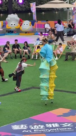 The video shows a performance with two people on stilts, accompanied by a clown on the ground. The stilt walkers have brightly colored pants that flare out in sections, one with blue and yellow and the other with blue, pink, and white. The clown is wearing a brown jacket, patchwork pants, and a hat. They are performing on a grassy area with a patterned stage at the bottom of the frame. Behind them, there is a seated audience in lawn chairs, a small pond, and two cartoon rock statues. The overall environment suggests an outdoor arts festival or public performance. There are several flags and banners visible in the background. The performers interact with each other, and the clown gestures with their hat.