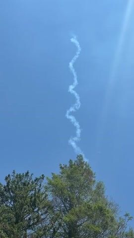 The video shows a bright blue sky with a contrail shaped like a zigzag running from the top of a tree towards the upper part of the frame. The top of two green trees are visible at the bottom of the frame. The sun is shining brightly.