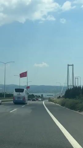 The camera records from inside a moving vehicle approaching a suspension bridge on a highway. The sky is blue with scattered clouds. Turkish flags are displayed along the road. The bridge's structure is visible in the distance. Other vehicles, including a bus and several cars, are on the road.