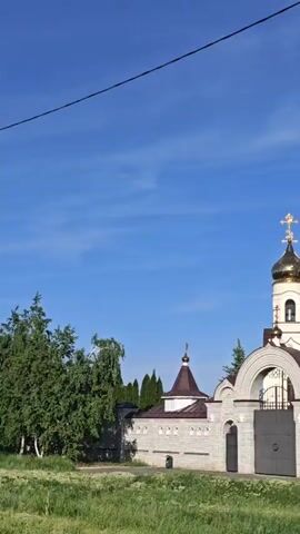 The video shows the exterior of a large, ornate church with multiple golden domes topped with crosses. The building is constructed of light-colored stone and has several arched windows. A brick wall surrounds the church grounds, with an arched gate at the entrance. The sky is a bright blue with some clouds. The surrounding area appears to be grassy, with some trees. The camera pans to show more of the wall around the church.