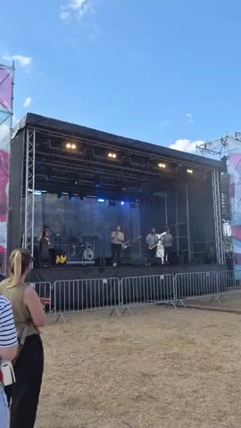 A band is performing on an outdoor stage. The stage is elevated and covered by a black canopy. Several band members are visible on stage, including a singer, a guitarist, and a drummer. Lights are set up on the stage's rigging. In the foreground, two women are standing, facing the stage. They are in a grassy, outdoor area, with a blue sky with clouds visible above. A fence separates the audience area from the stage. There are also some decorative structures visible on either side of the stage. The overall impression is that of an outdoor music event or festival.