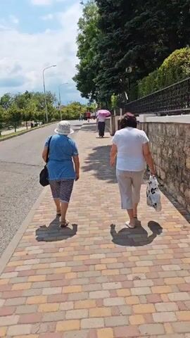 Two women are walking along a brick-paved sidewalk on a sunny day. The woman in the foreground on the left is wearing a blue denim top, patterned capri pants, and a white sun hat. She has a dark shoulder bag. The woman on the right is wearing a white t-shirt and light-colored capri pants. She is carrying a white plastic bag with black lettering. Both women are walking away from the camera.

In the background, another person is walking with a pink umbrella. The sidewalk is lined with a stone wall on the right, and a street with some trees and a lamppost on the left. Further in the distance, more trees and buildings are visible. The sky is blue with some scattered clouds. The lighting suggests it is daytime, likely late morning or early afternoon, given the length and direction of the shadows. The overall scene depicts a casual stroll in what appears to be a town or city environment, possibly a park or a promenade, given the well-maintained sidewalk and landscaping. The presence of the sun hats and umbrella suggests warm weather. The people appear to be relaxed as they walk. No visible text beyond the lettering on the plastic bag. The location context is Pyatigorsk, Russia.