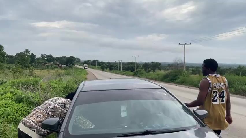 The video shows a scene beside a rural road in Oduape, Nigeria. A dark-colored car is parked on the shoulder of the road, its driver's side door open. A man wearing a yellow and white basketball jersey with the number 24 on it stands by the open door, looking towards the road. Another person, partially visible, is leaning into the car from the passenger side, seemingly retrieving something. The surrounding environment is lush and green, with dense vegetation and trees on both sides of the road. In the distance, a truck is visible driving down the road, and scattered buildings and power lines suggest a rural community. The sky is overcast with dramatic, cloudy formations, indicating a possibly impending rain or just a cloudy day. The overall impression is a moment paused on a journey, perhaps a brief stop for a personal matter or to attend to something in the car. The presence of the "PRESS" sticker on the car's windshield might suggest the occupants are journalists or involved in media work, although their current activity appears to be personal. The time of day appears to be daytime, given the ambient light.