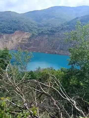 This video clip captures a scenic view of a bright turquoise lake nestled within a valley, surrounded by lush green mountains and a partially cleared mountainside. The foreground is dominated by the branches of trees, some bare and others covered in leaves, creating a natural framing effect for the broader landscape.

The lake itself is the central subject, its vivid blue color contrasting sharply with the earthy tones of the exposed rock face of the mountain behind it. This mountainside appears to have been subject to excavation or natural erosion, revealing distinct layers of soil and rock. The slopes of the surrounding mountains are densely forested, indicating a natural, possibly remote, environment.

The sky above is partially cloudy, suggesting it is daytime, likely late morning or afternoon. The overall impression is one of natural beauty and tranquility, with the vibrant lake and verdant mountains offering a picturesque landscape. The presence of bare branches in the foreground might suggest a season transitioning into or out of full foliage, or simply a particular type of vegetation. No people or specific objects are visible, placing the focus entirely on the natural scenery.

The location context of Panagyurski kolonii, Bulgaria, places this scene in a mountainous region known for its natural landscapes. The image evokes a sense of peace and awe at the grandeur of nature.