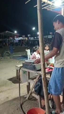 A nighttime outdoor scene shows a man wearing a light-colored shirt and patterned blue shorts standing near a food stall. In front of him is a metal cooking surface with a large round pan on top. Two young girls are visible near the cooking surface, one in a white outfit and the other in a plaid shirt. In the background, there are people gathered, with lights and banners suggesting an event or market. The ground appears to be dirt. A black plastic bag hangs from the stall's structure.