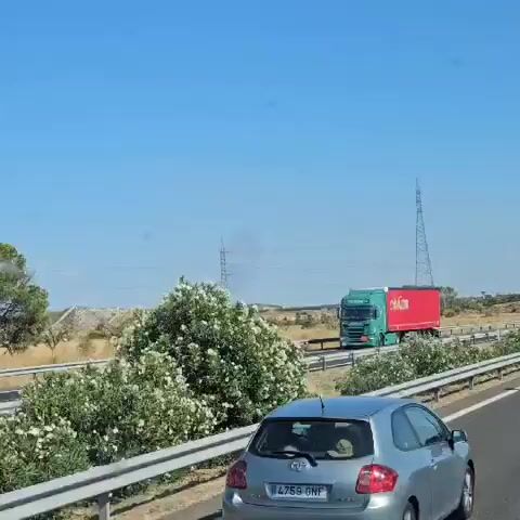 The video shows a highway scene on a clear, sunny day. A silver car is in the foreground, driving on the right lane. A large red semi-trailer truck with the word "CALZADOS" visible on its side is passing the car on the left lane. In the background, there are rolling hills, dry vegetation, and power lines. A plume of dark smoke is visible rising from the hills in the distance.