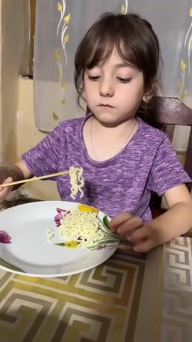 A young girl with brown hair and brown eyes is sitting at a table and eating noodles with chopsticks. She is wearing a purple shirt. The table has a patterned tablecloth. The noodles are on a white plate with a floral design. The girl is looking at the camera. The background is blurry and appears to be a room with a window and curtains. The girl picks up a strand of noodles with her chopsticks and brings it to her mouth, then chews. She then lowers the chopsticks and looks at the camera again.