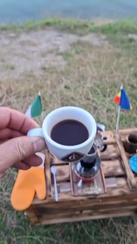 A person's hand holds a white espresso cup filled with dark coffee. In the background, a body of water reflects the sky, which transitions from the muted colors of early morning or late evening to brighter hues as the scene progresses. The foreground features dry grass and a wooden crate with camping equipment, including a portable stove and flags. The camera slowly zooms out, revealing more of the serene waterside environment as the light changes.